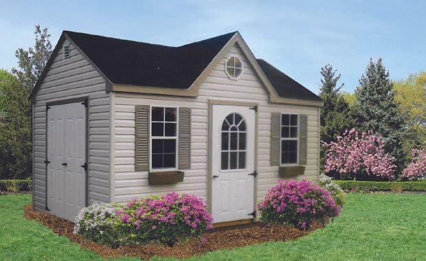 Tan shed with black roof, white door, and shutters. Surrounded by flowers and trees in a yard.