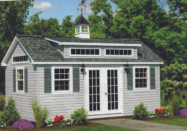 Gray shed with white trim, green shutters, French doors, and a cupola; surrounded by plants and trees.