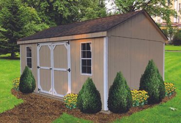 Tan shed with white trim, double doors, two windows, and dark roof, on a lawn with bushes and flowers.