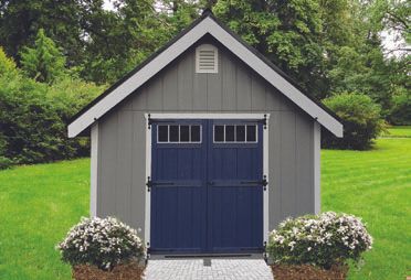 Gray and blue shed in a yard, with a gable roof, double blue doors, and flowers in front.