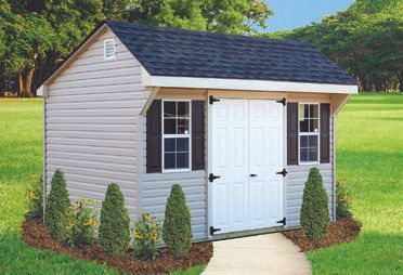 Tan shed with white doors, black shutters, and a dark roof, on a grassy lawn with trees.