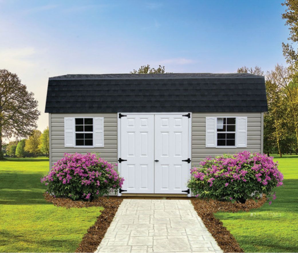 Tan storage shed with black roof, white doors and shutters, stone path, and purple bushes.