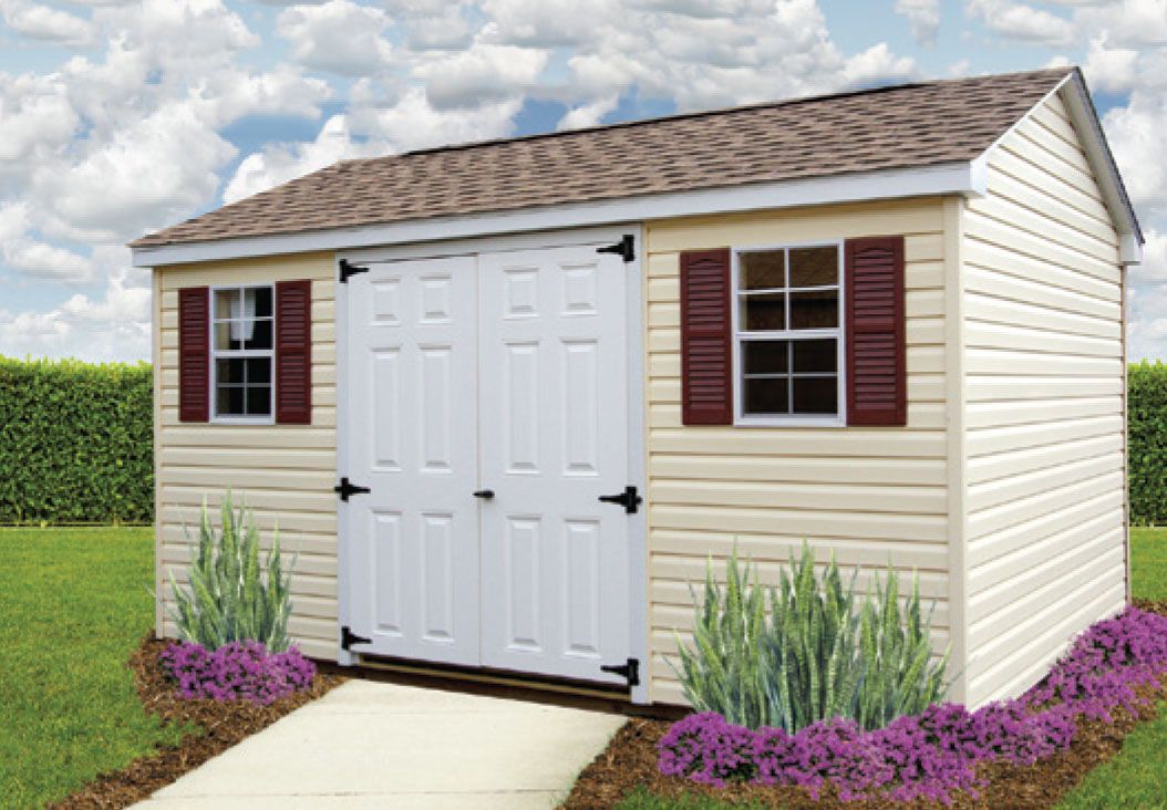 Tan shed with brown roof, white doors, and burgundy shutters on a lawn with bushes.