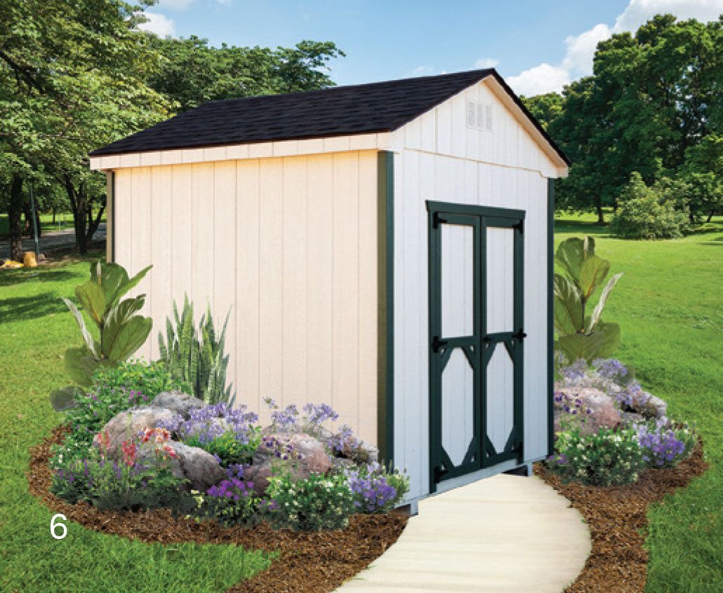 White and green shed with a black roof in a garden, flowers in front, a stone path leads to it.