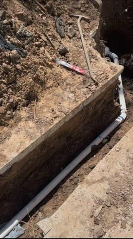 Trench with white PVC pipes laid along its side. A shovel and a saw are nearby. Dirt surrounds the trench.