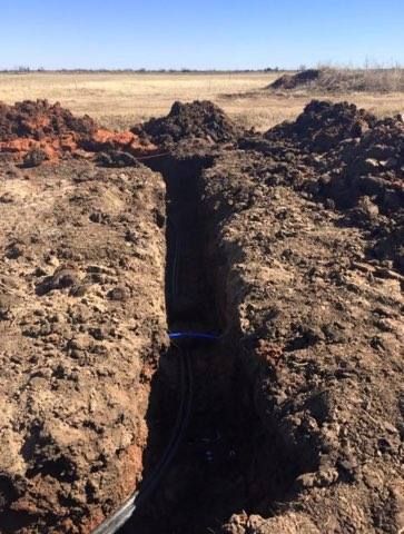 Trench in brown soil with exposed black and blue pipes, set in a field under a clear blue sky.