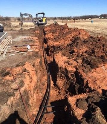 Trench with white PVC pipes laid along its side. A shovel and a saw are nearby. Dirt surrounds the trench.