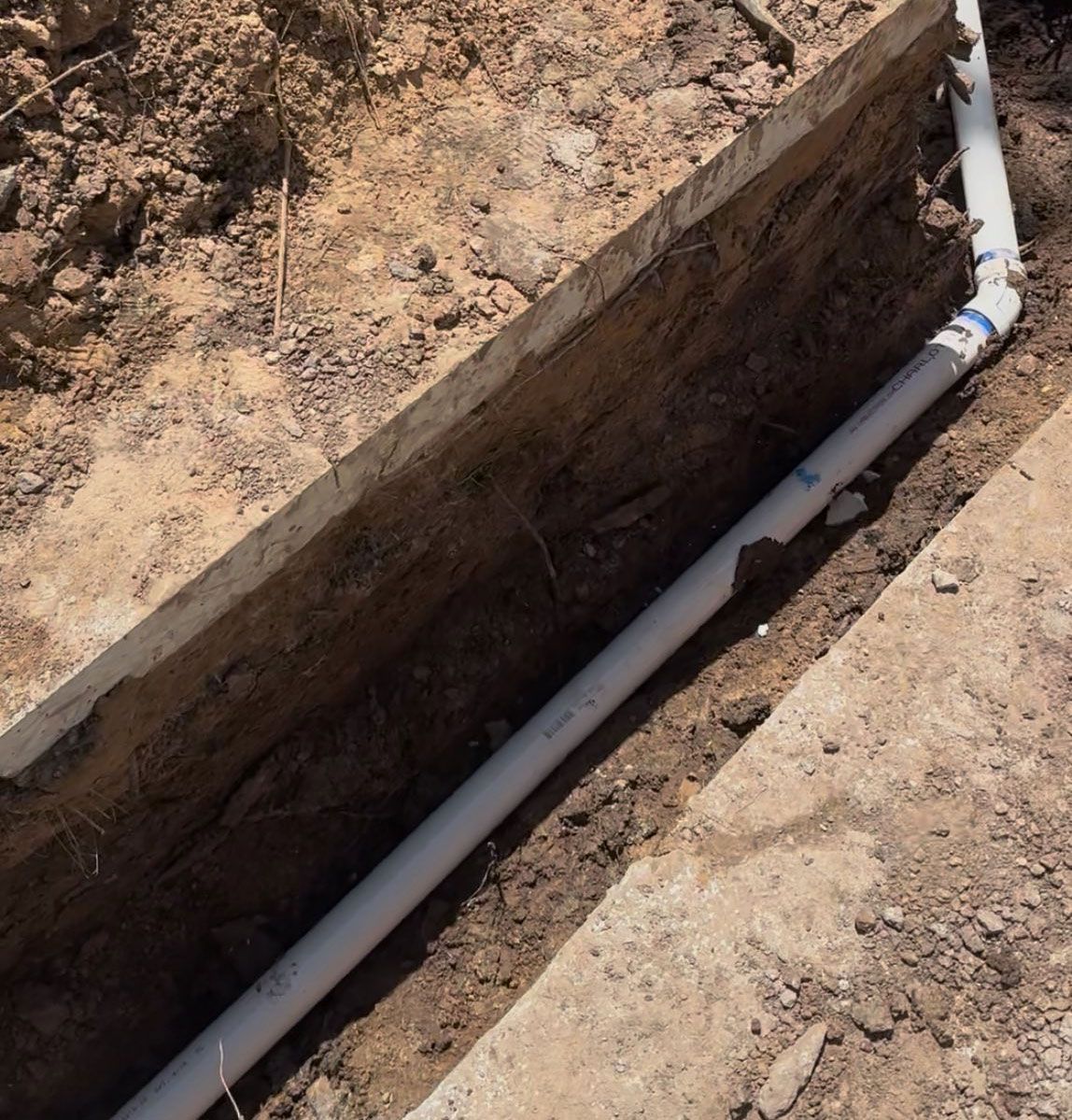 White PVC pipe laid in a trench, dirt and concrete surrounding it, outdoors.