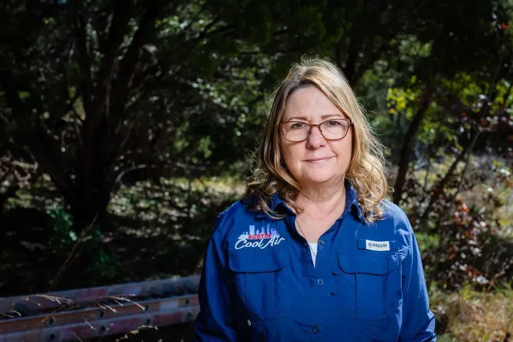 Woman wearing glasses and a blue shirt stands outside near greenery.