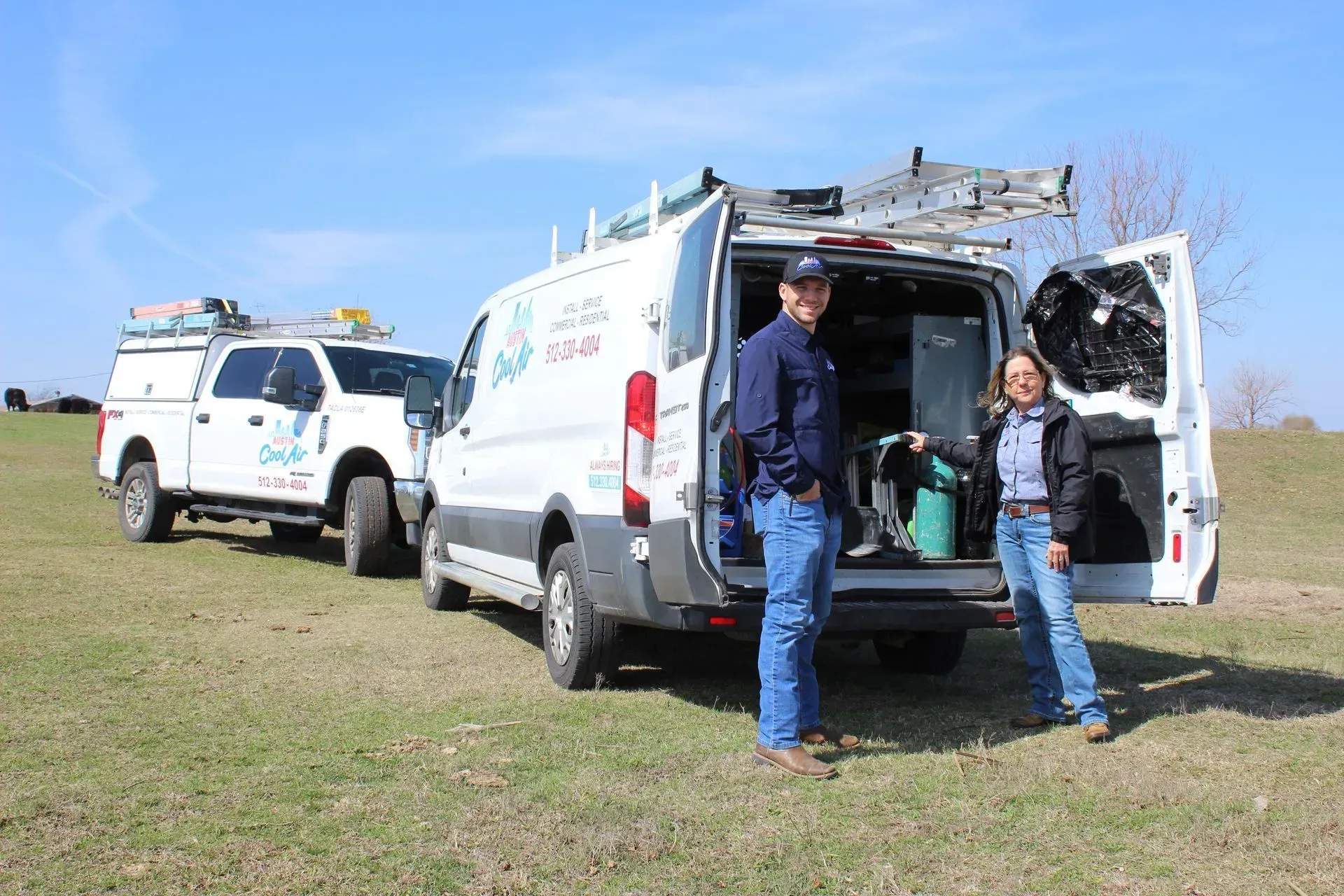 Two people stand near an open-doored work van and truck in a grassy field. Blue sky.