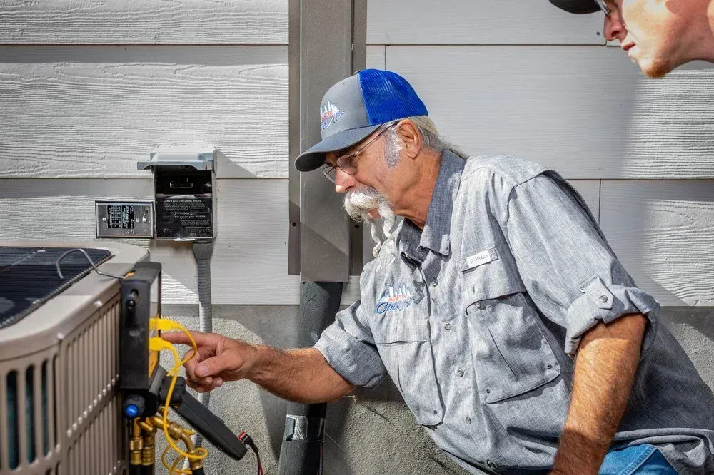 Man in hat pointing at AC unit with another man. Outdoors, next to house.