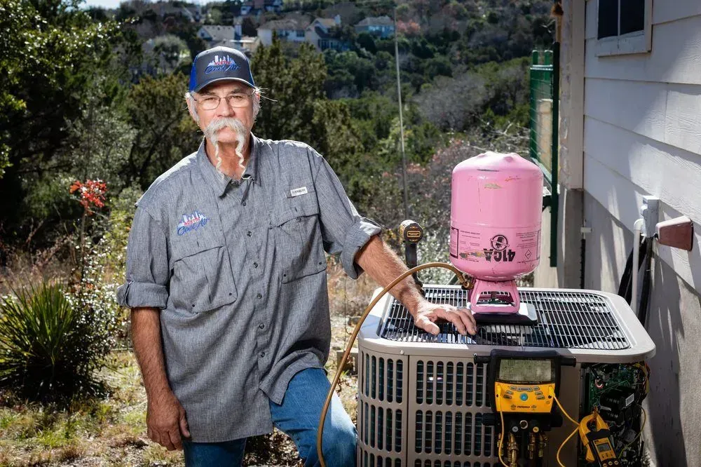 HVAC technician standing next to an air conditioning unit, pink tank on top. Outdoors, sunny.