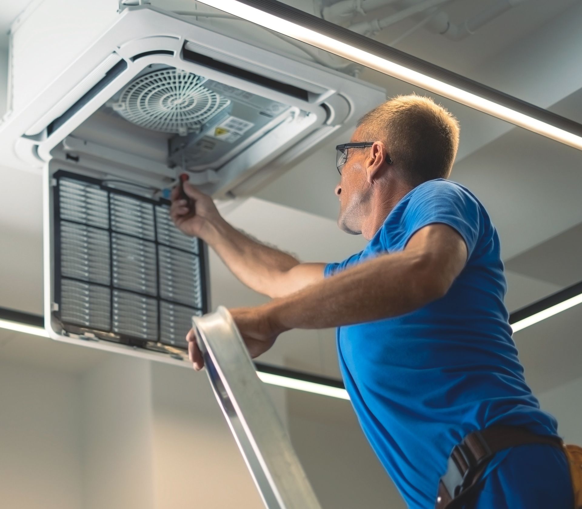 Man on a ladder servicing a ceiling air conditioning unit, removing a filter.