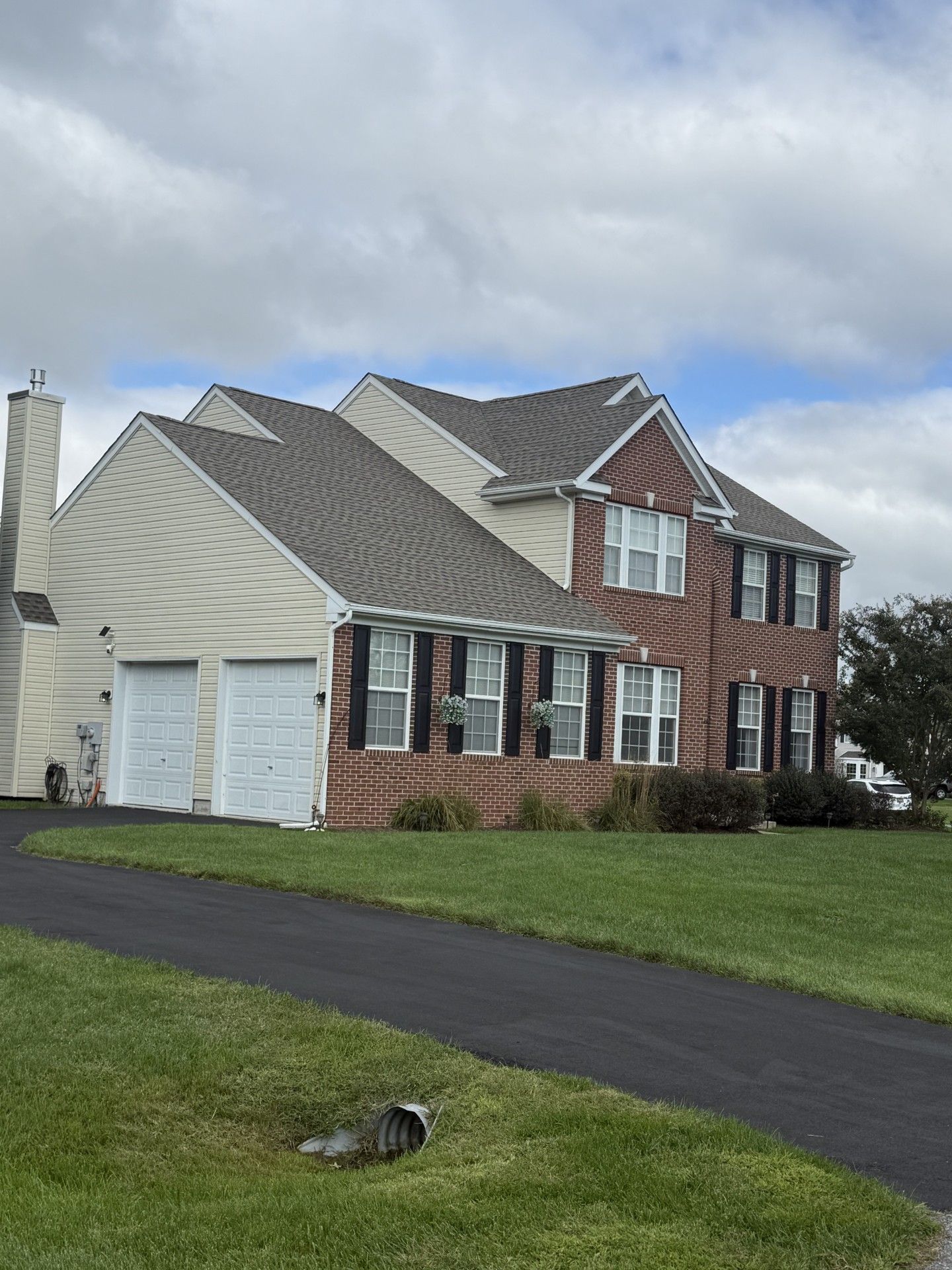 Two-story house with red brick and beige siding, black shutters, two-car garage, and a paved driveway.