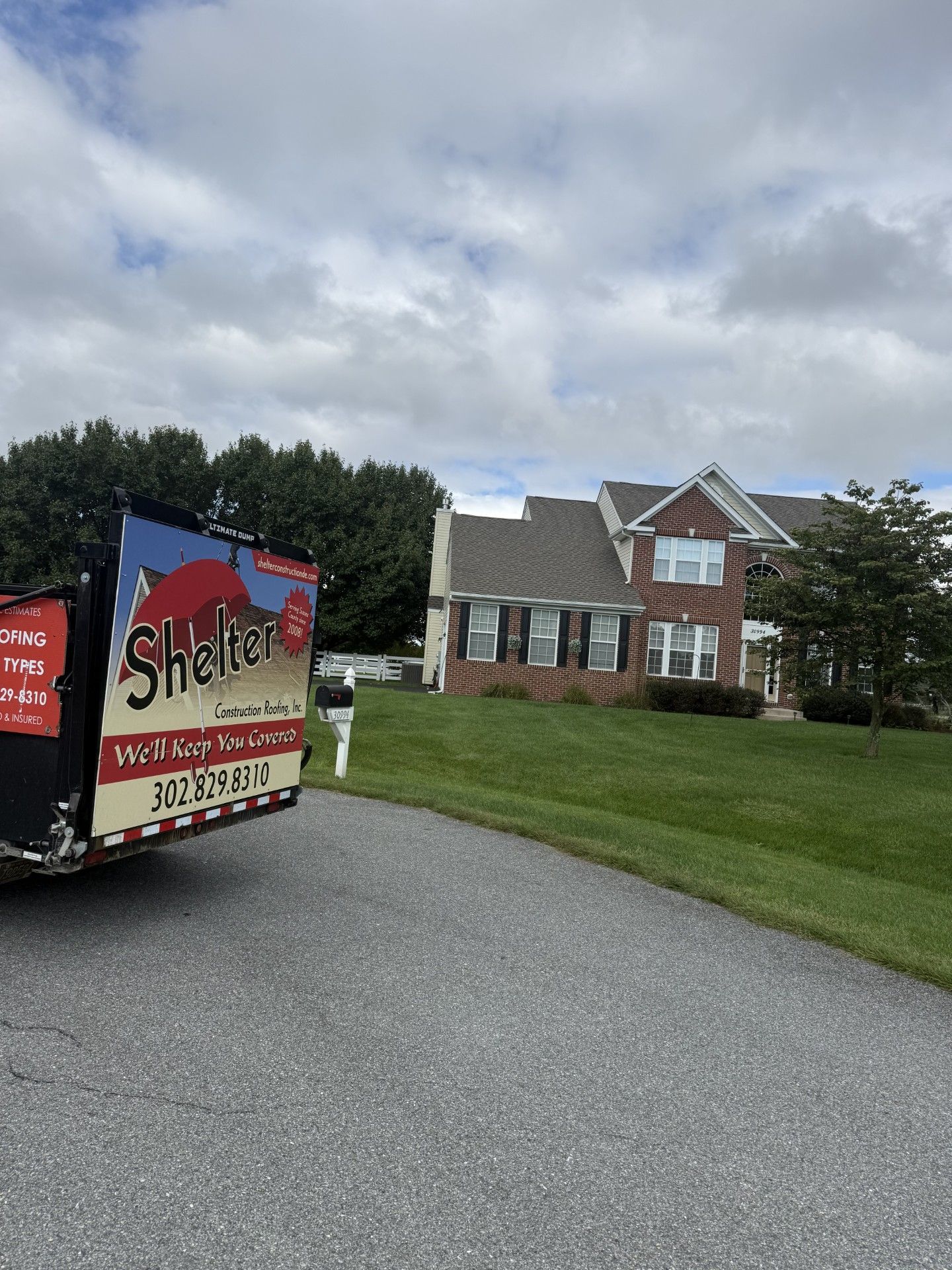 A red, blue, and white Shelter Solutions container in a driveway next to a two-story brick house under a cloudy sky.