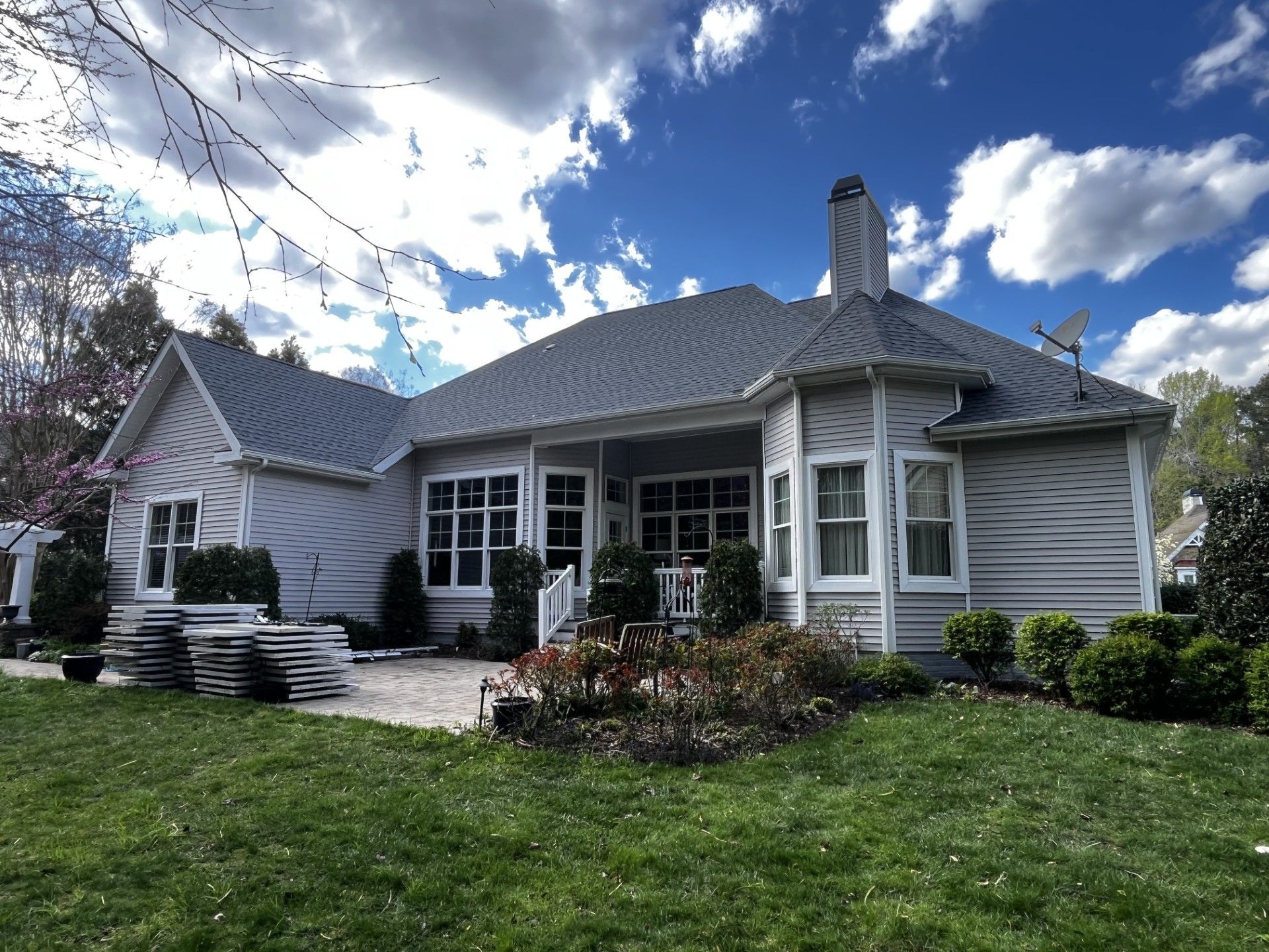 Backyard view of a light gray house with a gray roof, a brick patio, and a green lawn under a partly cloudy blue sky.