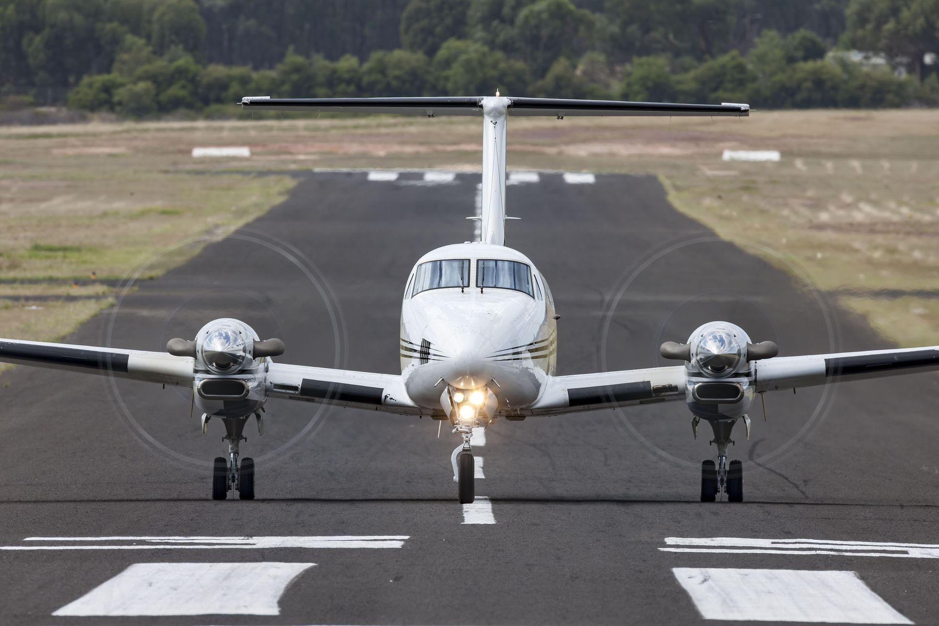 Twin-engine airplane on a runway, white and yellow with propellers, facing the viewer, ready for takeoff.