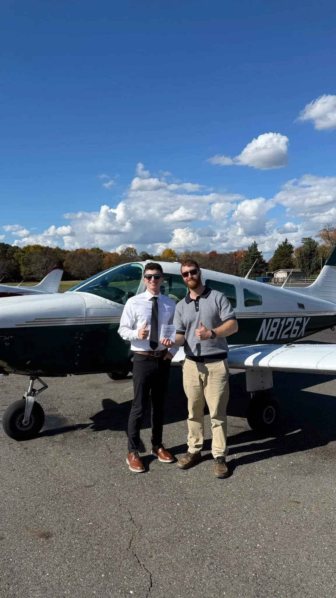 Two men stand in front of a small airplane on a sunny day. One holds a certificate, both give thumbs up.