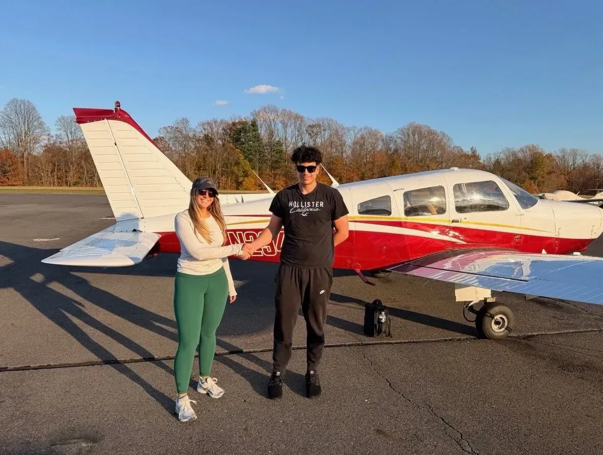 Two people shake hands in front of a small airplane on a tarmac, sunny day.