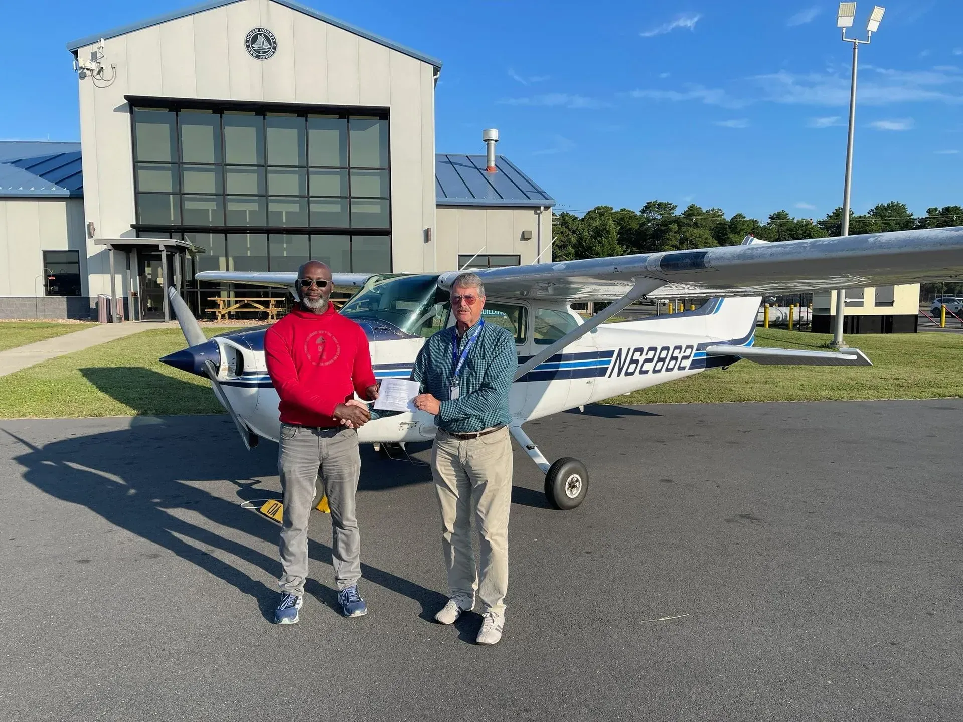 Two men stand in front of a small airplane near a hangar. One wears a red shirt, the other a patterned blue.