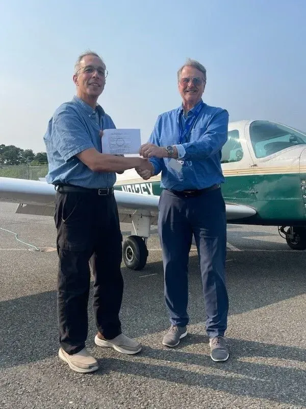 Two men shaking hands in front of a small airplane. One man holds a paper. Blue and green airplane. Sunny day.