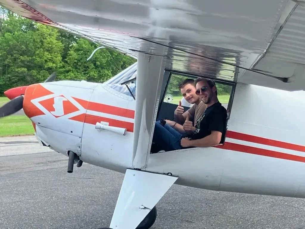 Two people smiling and giving thumbs up in a small airplane cockpit; exterior view.