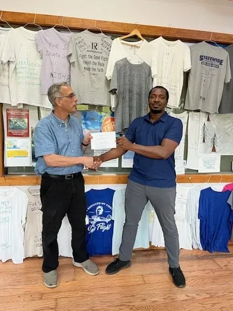 Two men shaking hands in front of a wall of t-shirts, indoors. One man wears a blue shirt and the other wears a dark blue shirt.
