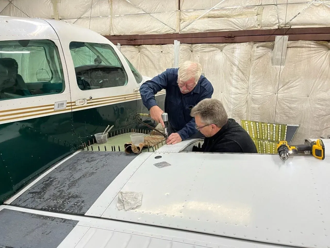 Two people working on the wing of a small airplane in a hangar, using tools.