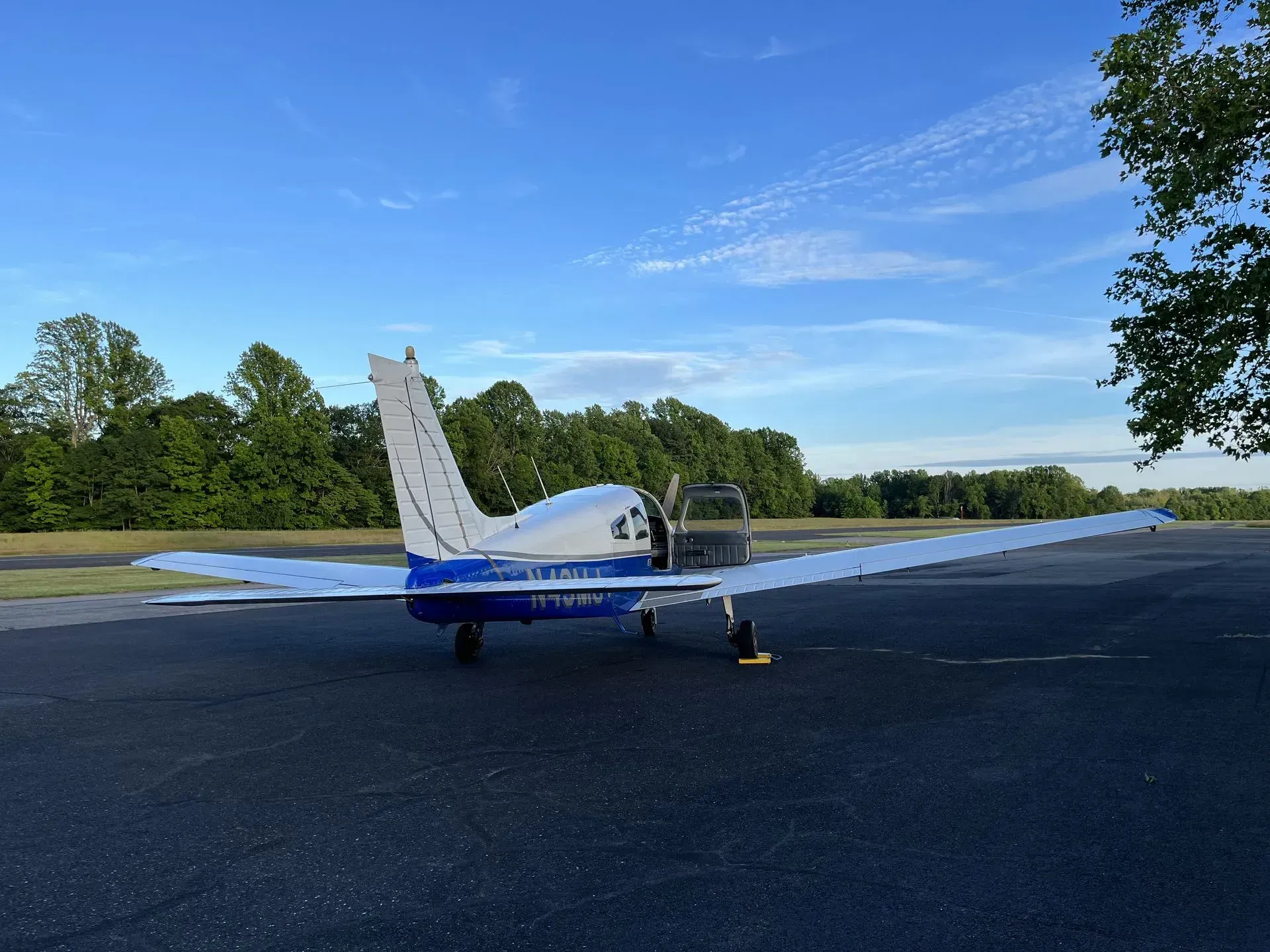 Small blue and white airplane parked on asphalt runway, trees in background, clear sky.