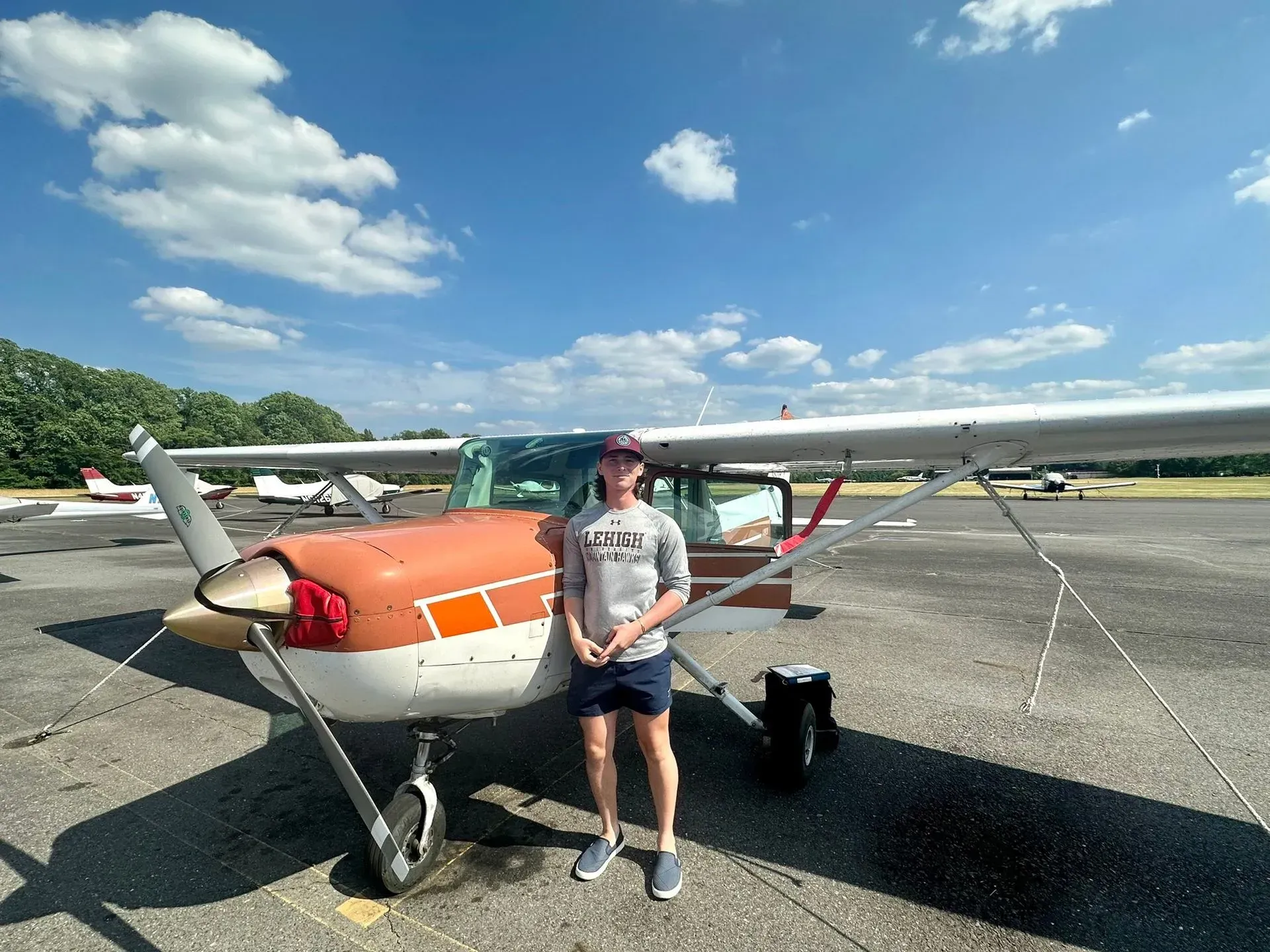 Man stands beside a small, orange and white airplane on a sunny airfield.