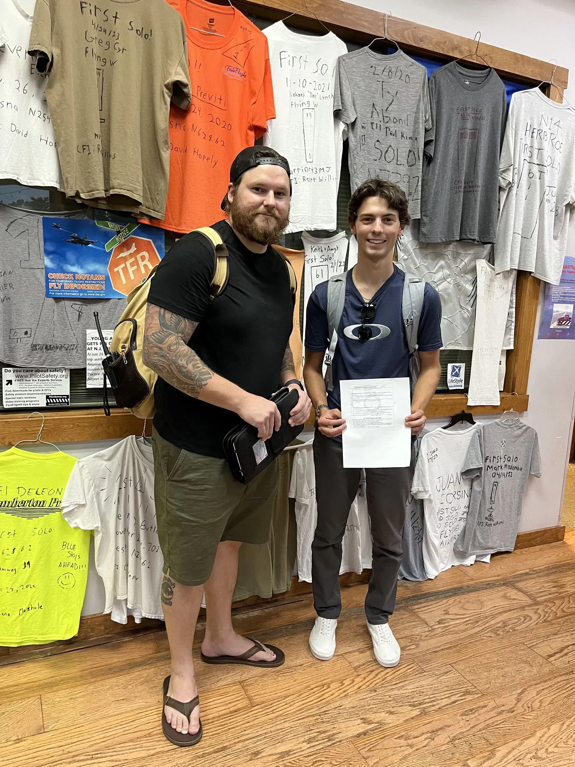 Two people in front of a wall of t-shirts. One man has tattoos and holds a camera. A younger person holds a paper.