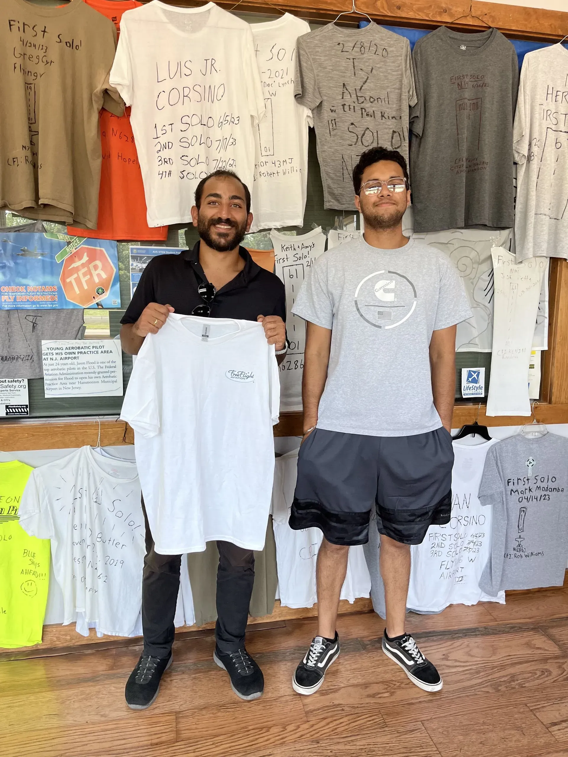 Two men stand in front of a wall of t-shirts. One holds a white tee, smiling; the other wears a gray tee, looking at the camera.
