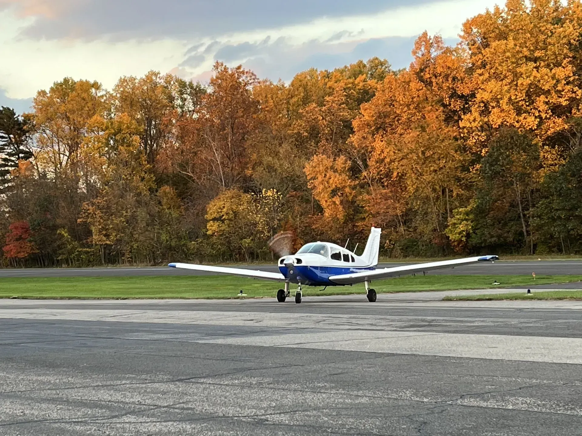 Small white and blue airplane on runway with autumn trees in background.