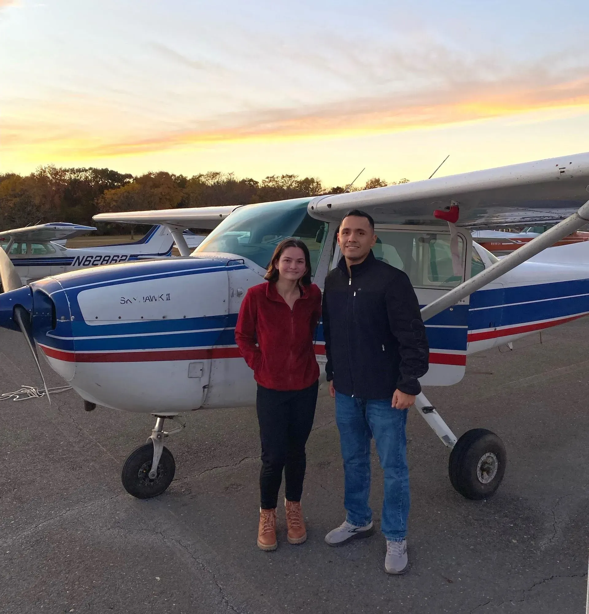 Two people stand in front of a small airplane on a tarmac at sunset.