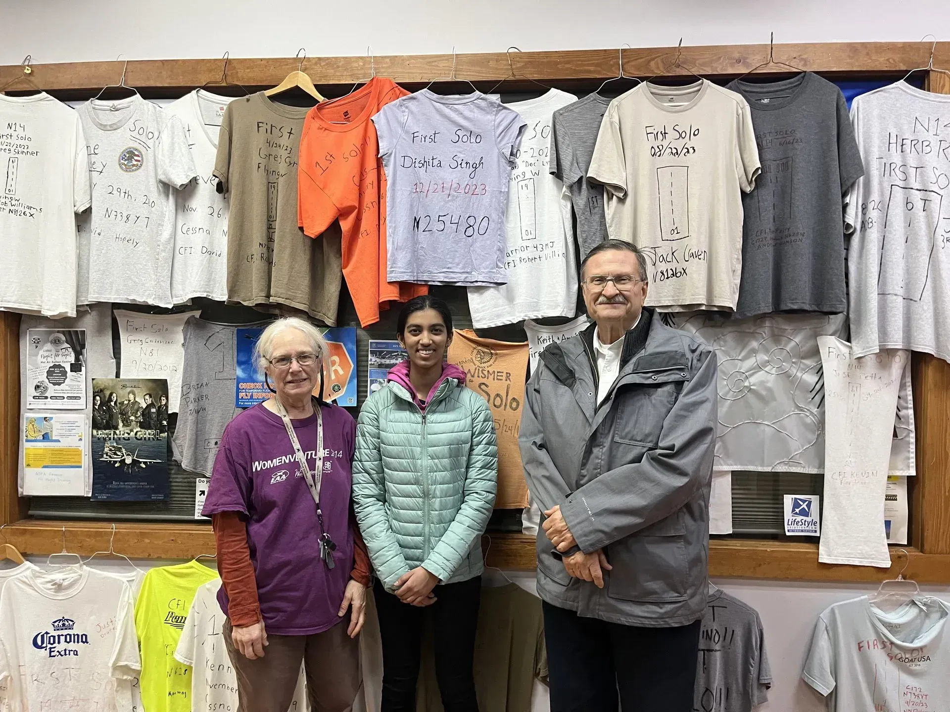 Three people stand in front of a wall of t-shirts. The wall is brown. The people are smiling.