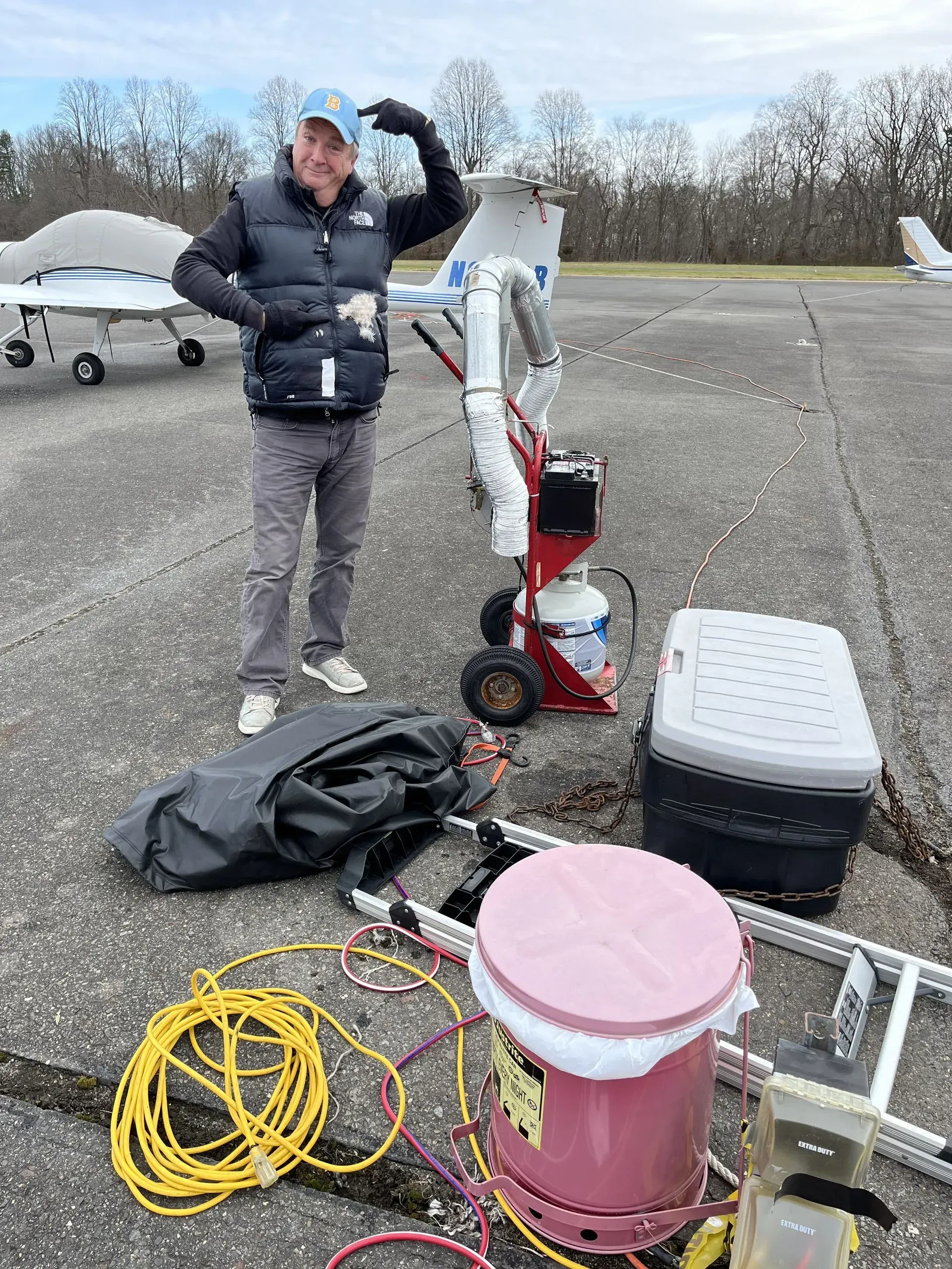 Man standing on an airstrip, pointing at his head; next to tools and equipment.