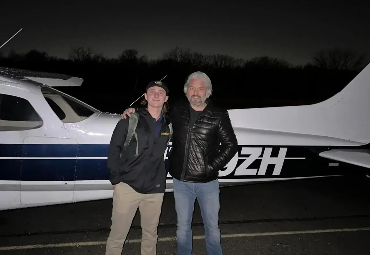 Two men stand by a small airplane at night. One has an arm around the other's shoulders.