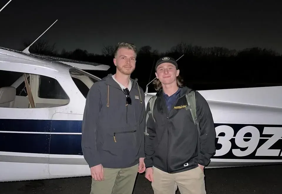 Two men stand in front of a small airplane at night. One wears a jacket, the other a hoodie.