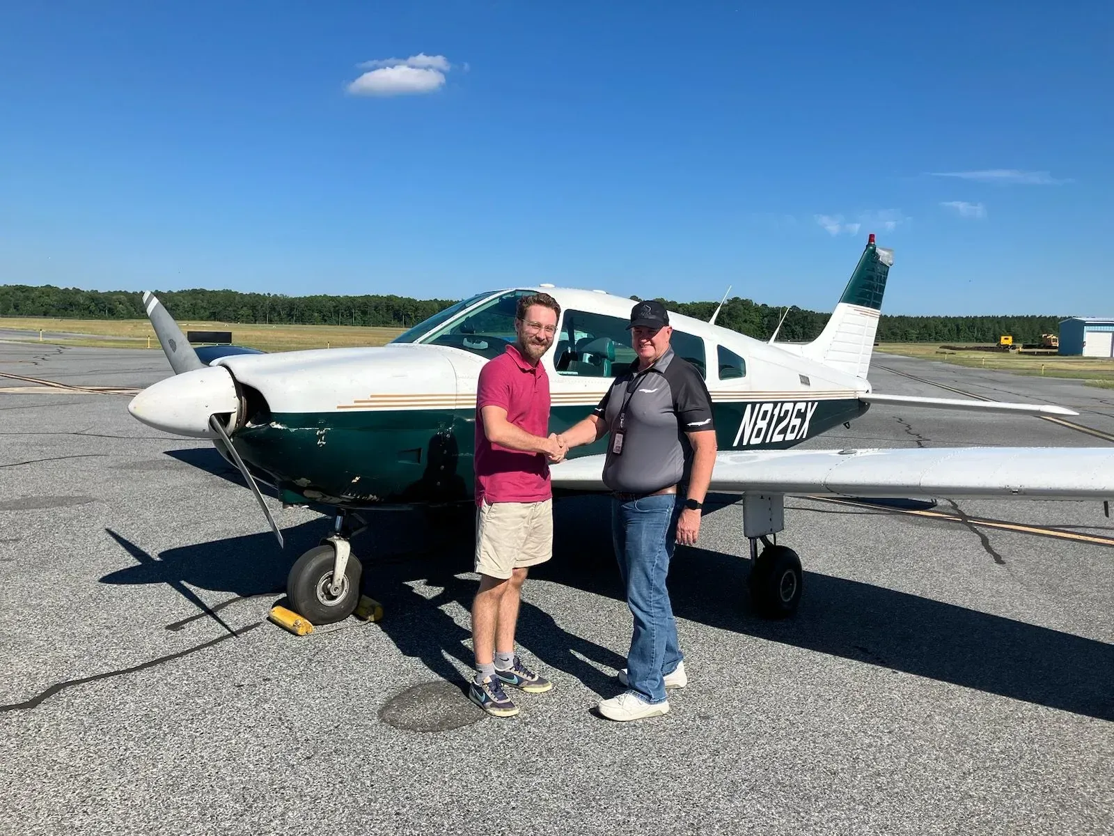 Two men shake hands in front of a green and white airplane on a tarmac under a blue sky.