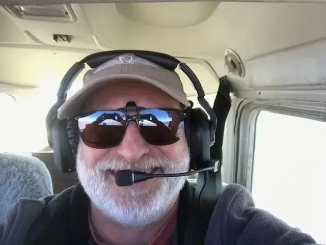 Man with a white beard wearing sunglasses, a headset, and a hat in an airplane cockpit; smiling.