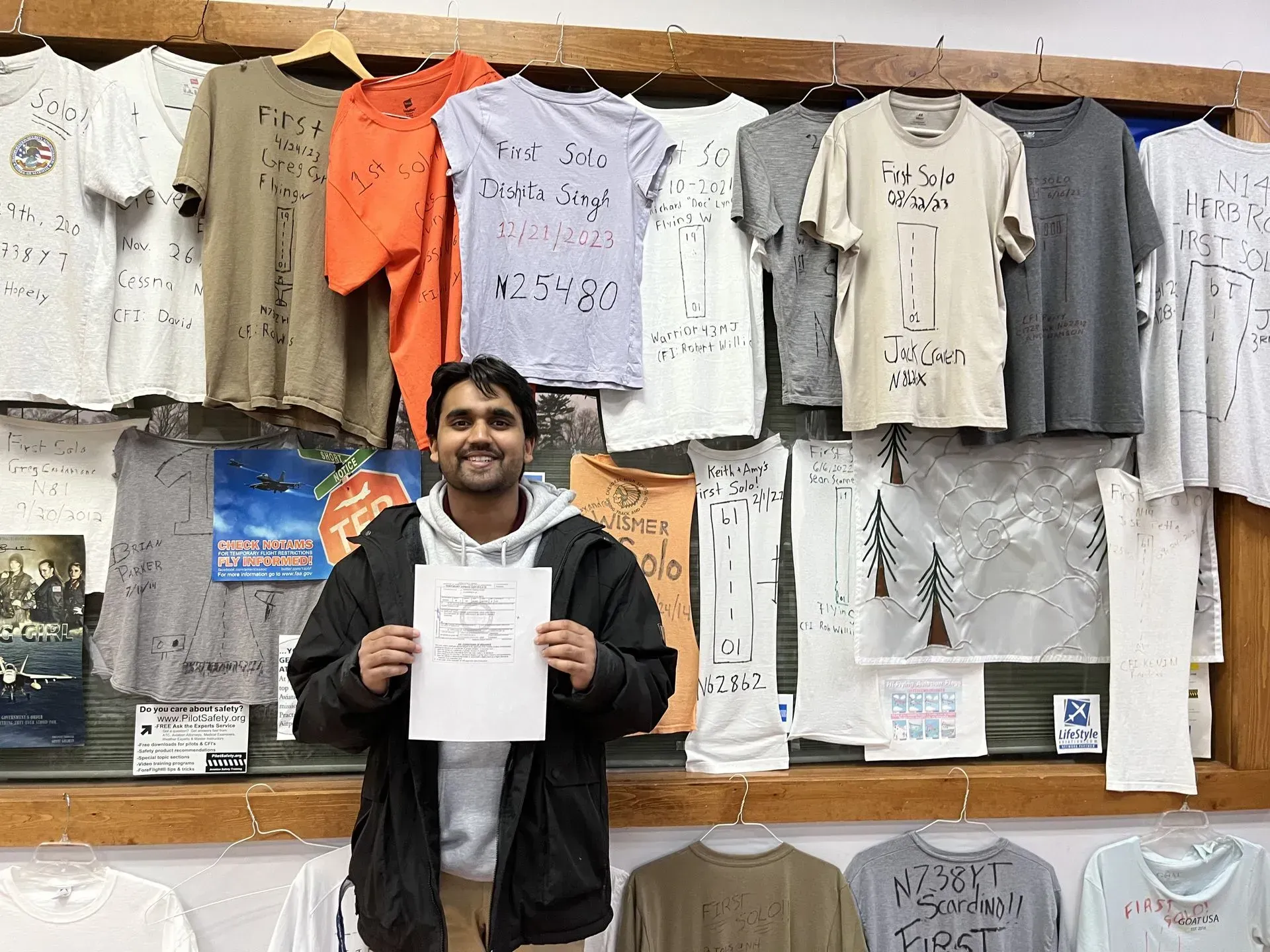 Person smiling, holding paper, in front of t-shirts hanging on wall.