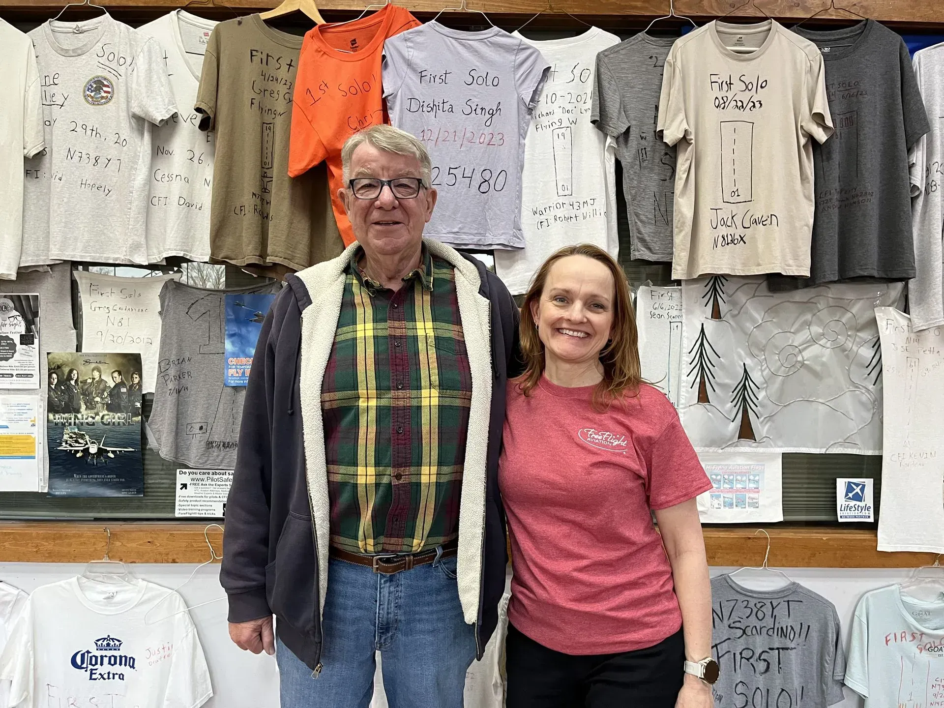 Man and woman standing in front of a wall of t-shirts. Man wears a plaid shirt, woman wears a pink shirt.