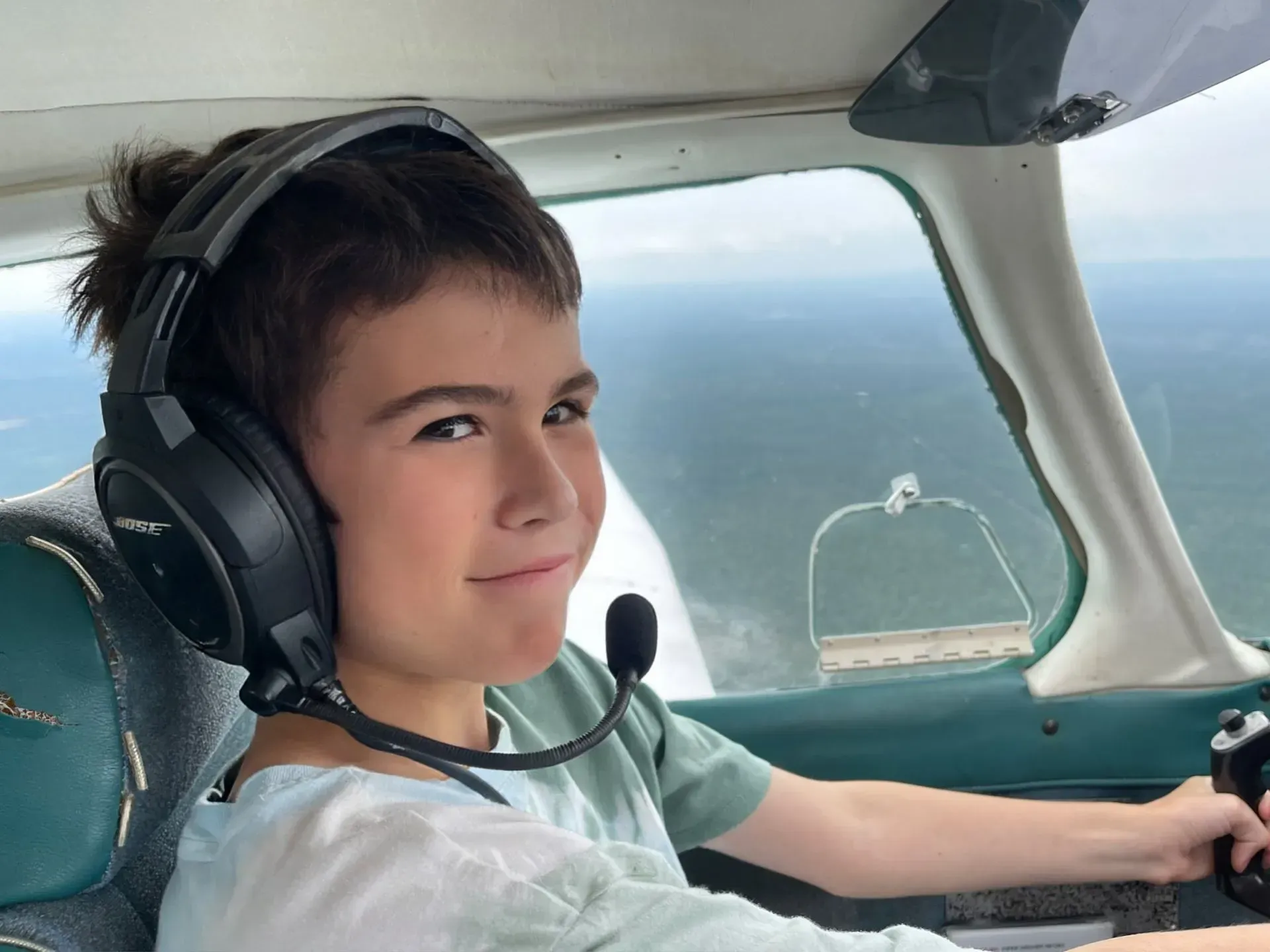 Young person wearing headset smiles while sitting in a cockpit. Ocean visible outside.