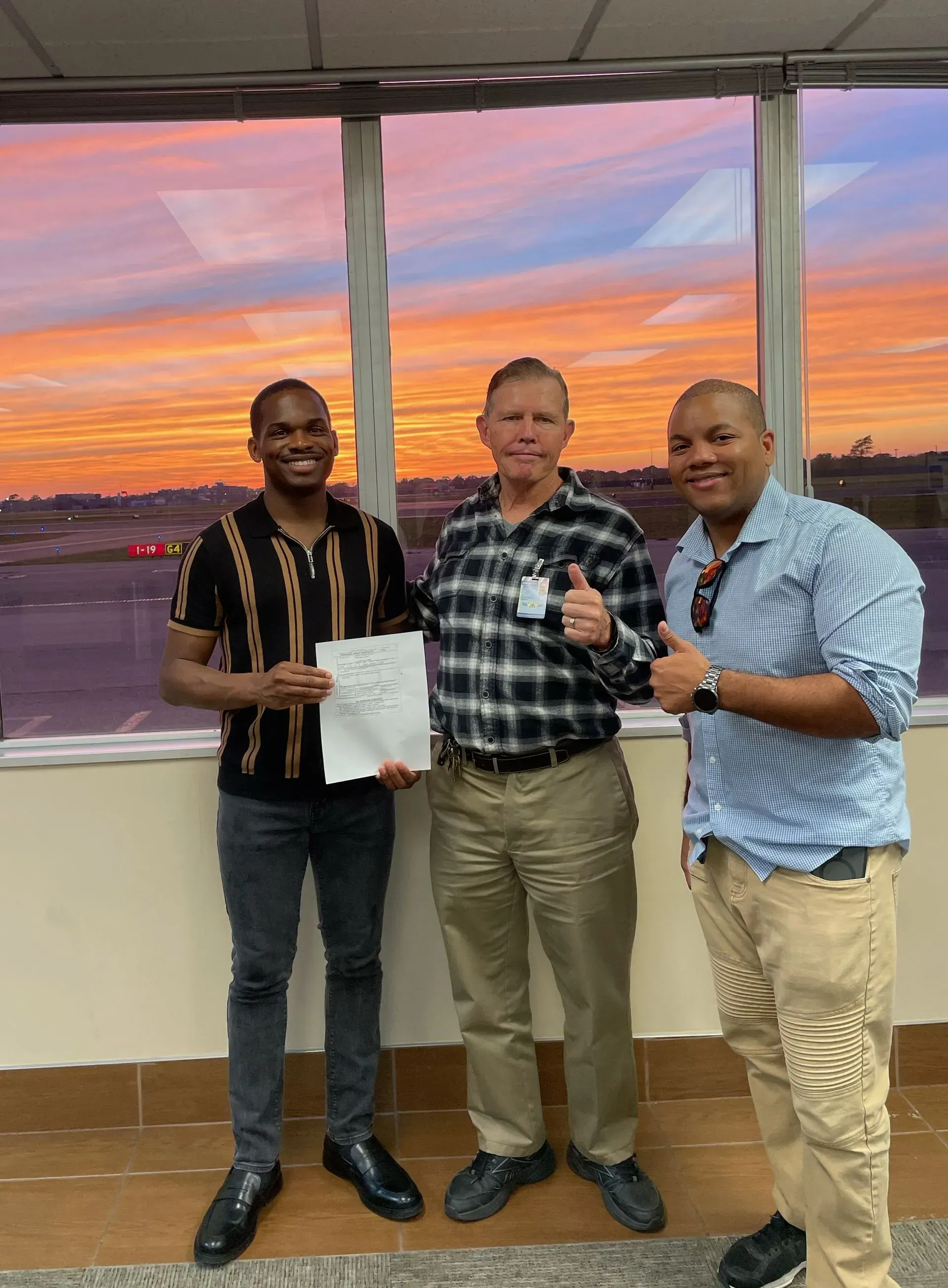 Three men smiling, thumbs up, by airport window with sunset view.