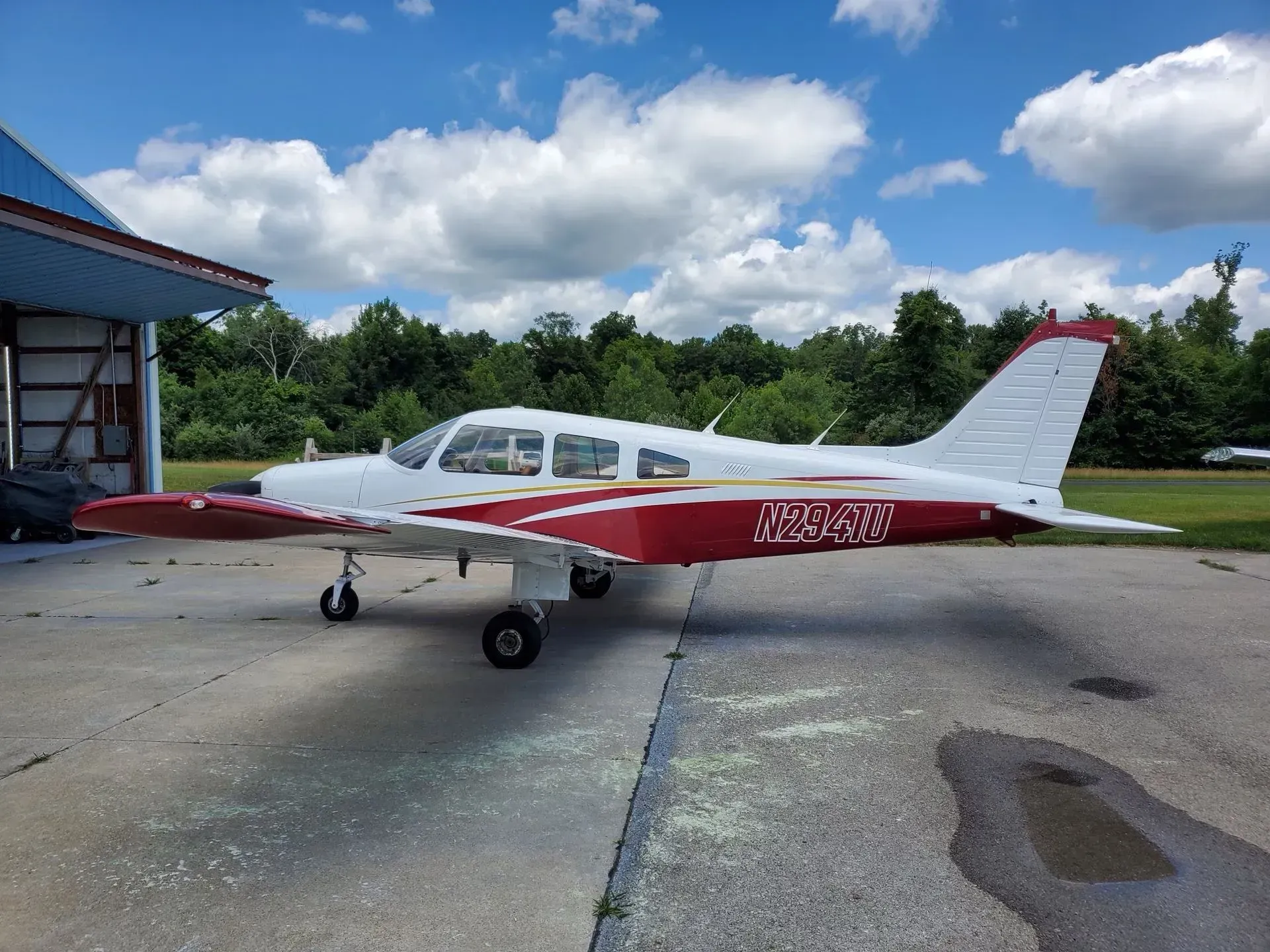 White and red single-engine airplane parked on a tarmac in front of a blue hangar, under a blue sky with clouds.