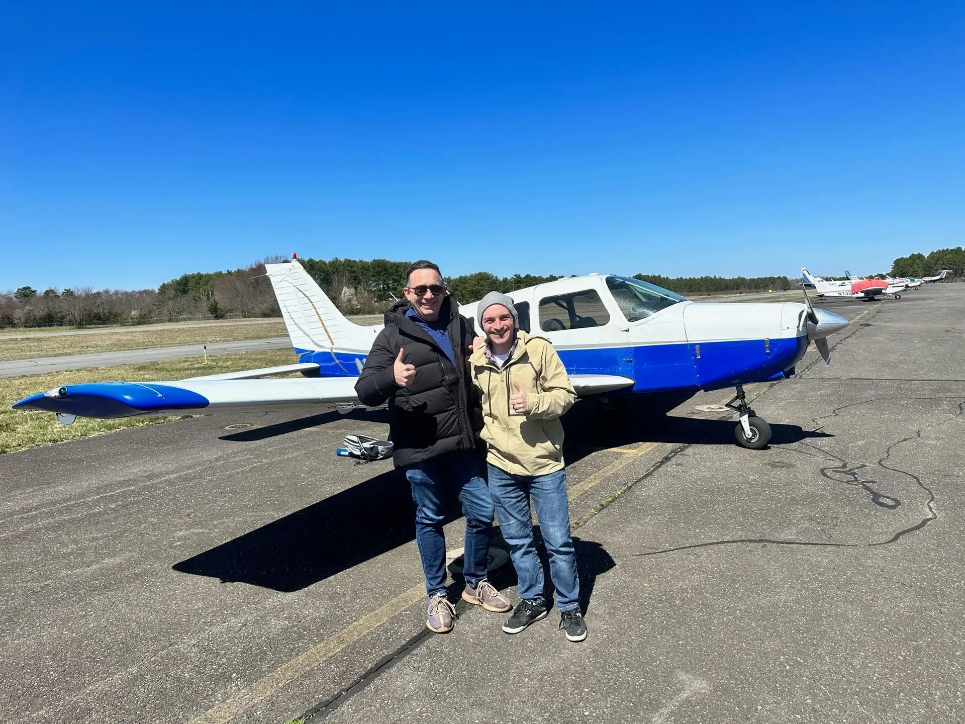 Two men standing in front of a blue and white airplane on a runway, sunny day. One man gives a thumbs up.