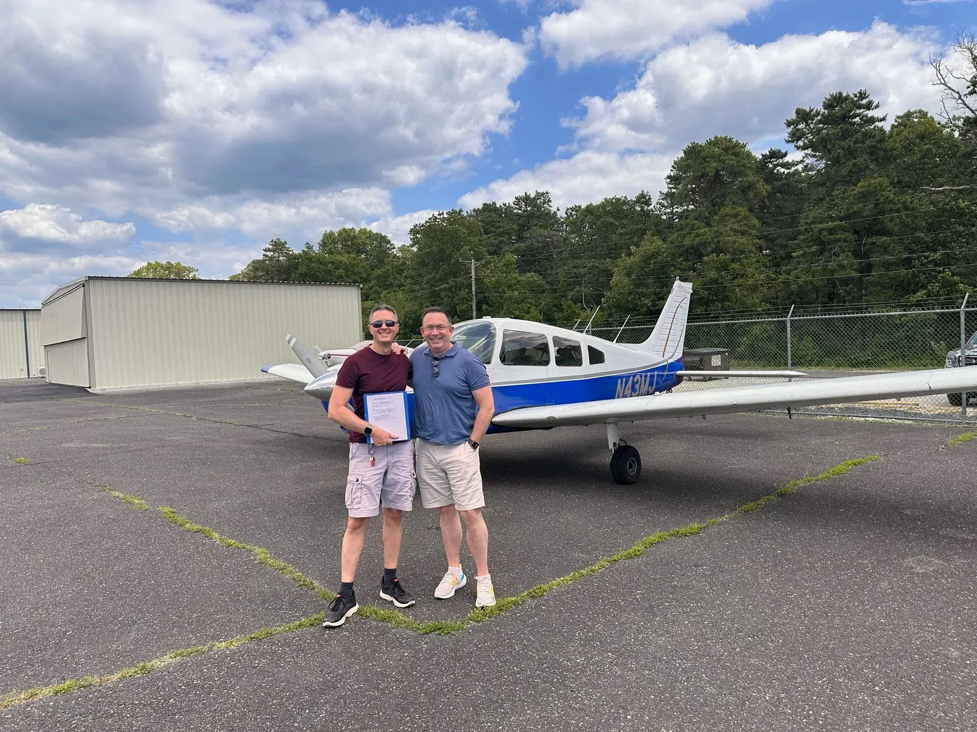 Two men stand by a small airplane, one holding a paper. Airplane is white and blue, outdoors on a tarmac.