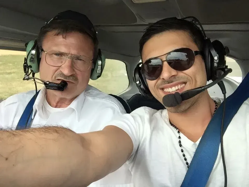 Two men in a small airplane cockpit, wearing headsets. One smiles, wearing sunglasses, the other has a serious expression.