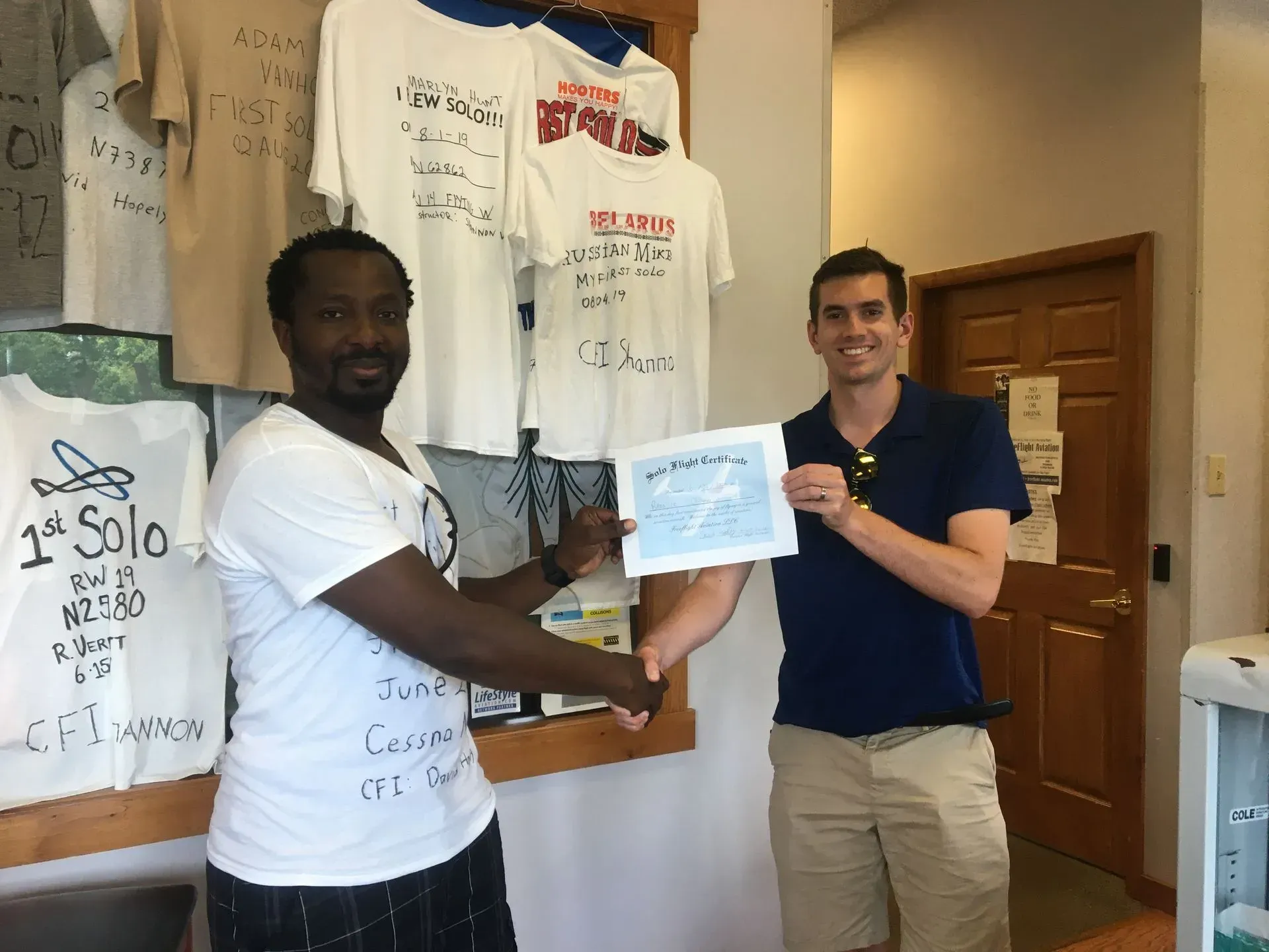 Two men shaking hands, one holding a certificate. T-shirts hang in the background. Indoor setting.