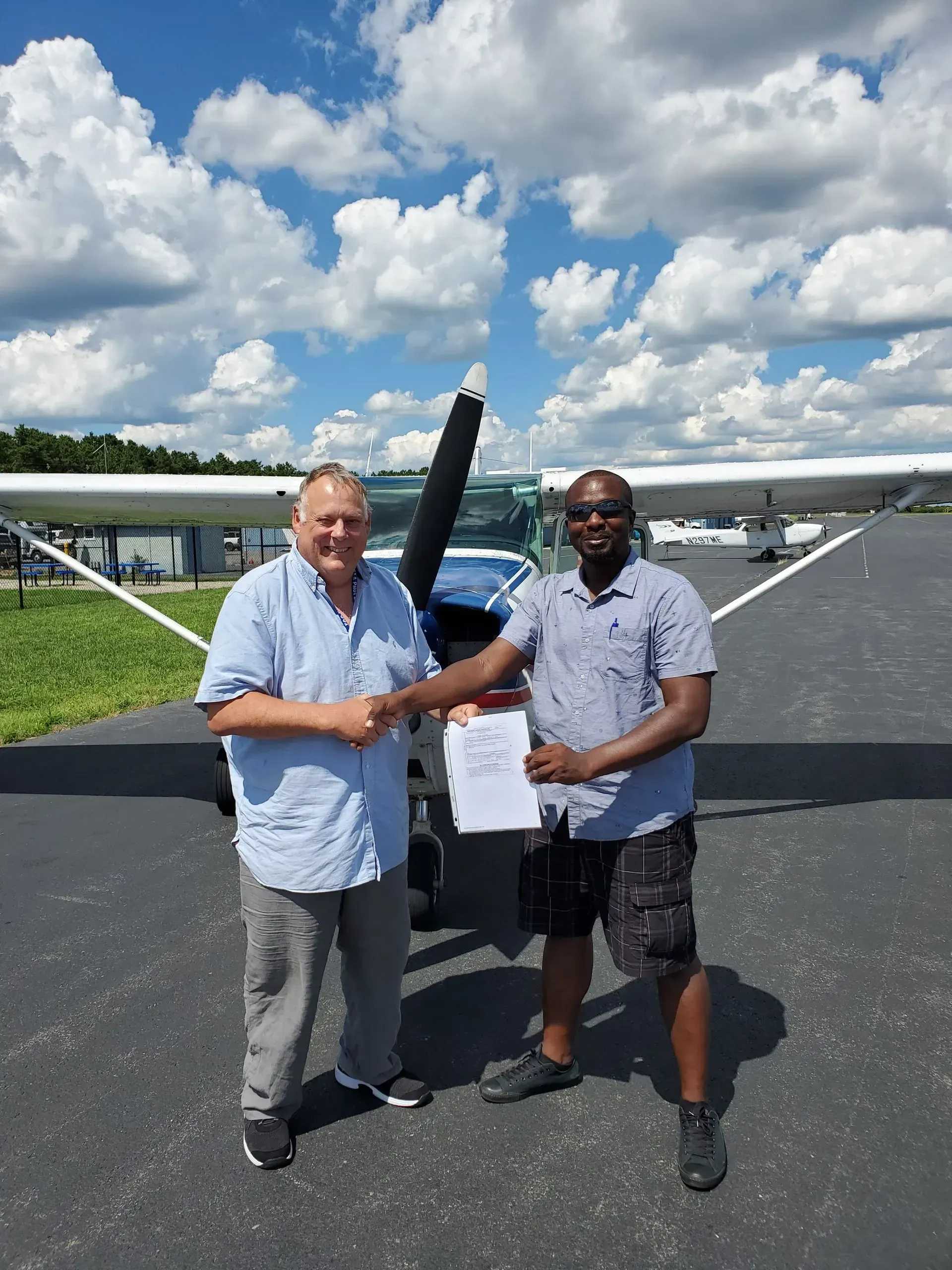 Two men shaking hands in front of a small airplane. Sunny day, blue sky with clouds.